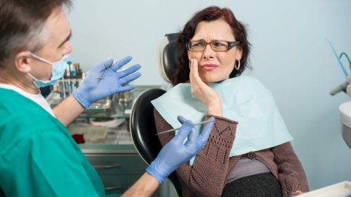 Woman feeling toothache, touching cheek with hand at dental clinic. Senior dentist trying to help. Dental care and health concept
